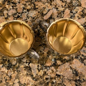 May include: Two gold-colored metal sugar bowls with fluted edges and silver handles. The bowls are empty and sitting on a brown and tan speckled surface.