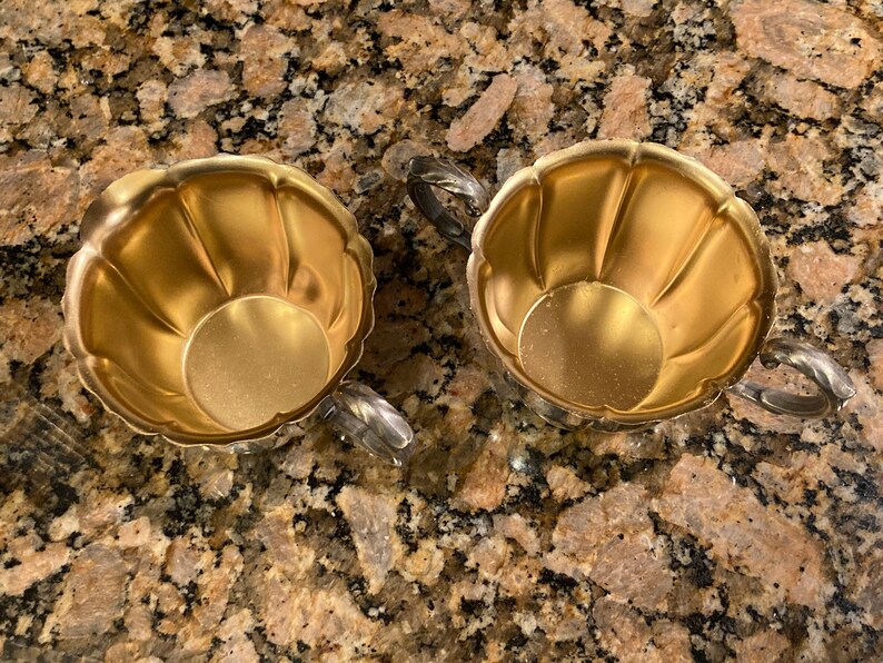 May include: Two gold-colored metal sugar bowls with fluted edges and silver handles. The bowls are empty and sitting on a brown and tan speckled surface.