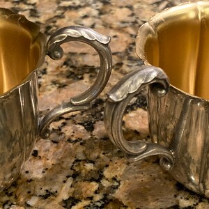 May include: Two silver sugar bowls with ornate handles and gold interiors. The bowls are sitting on a brown and white speckled countertop.