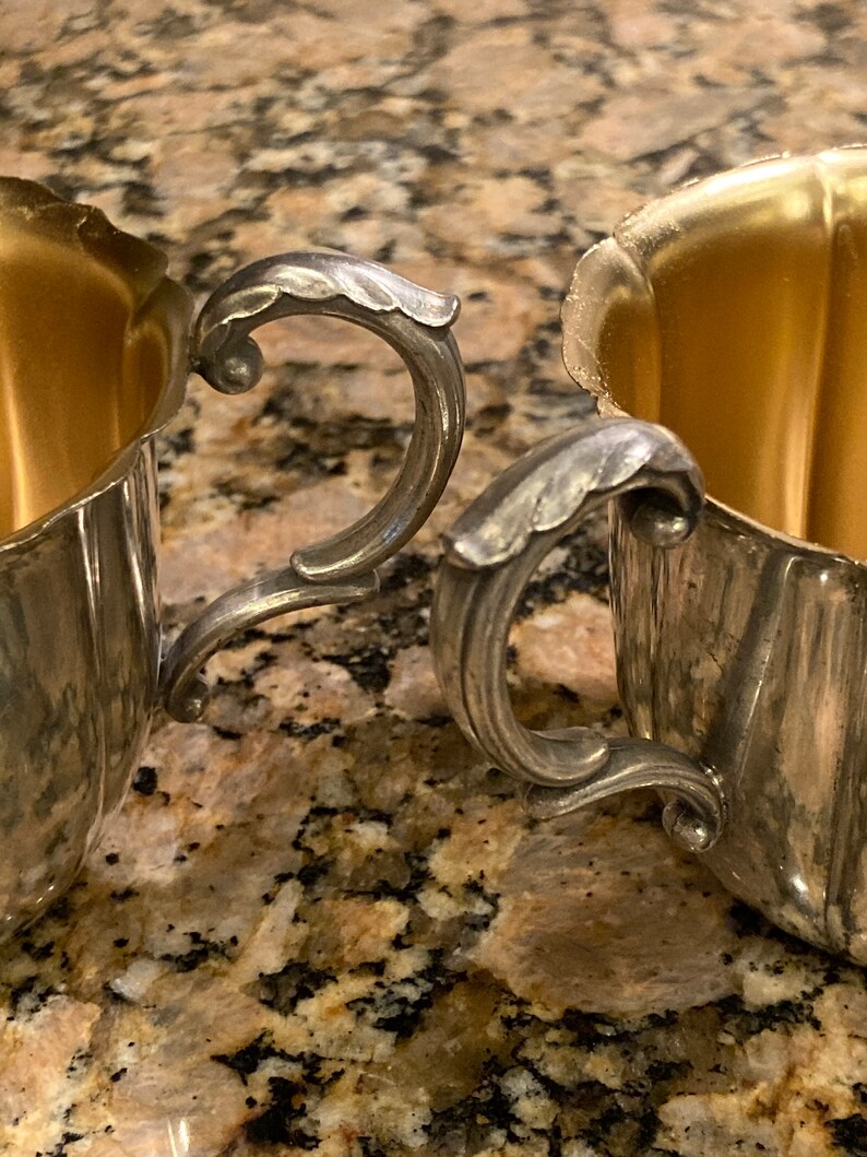 May include: Two silver sugar bowls with ornate handles and gold interiors. The bowls are sitting on a brown and white speckled countertop.