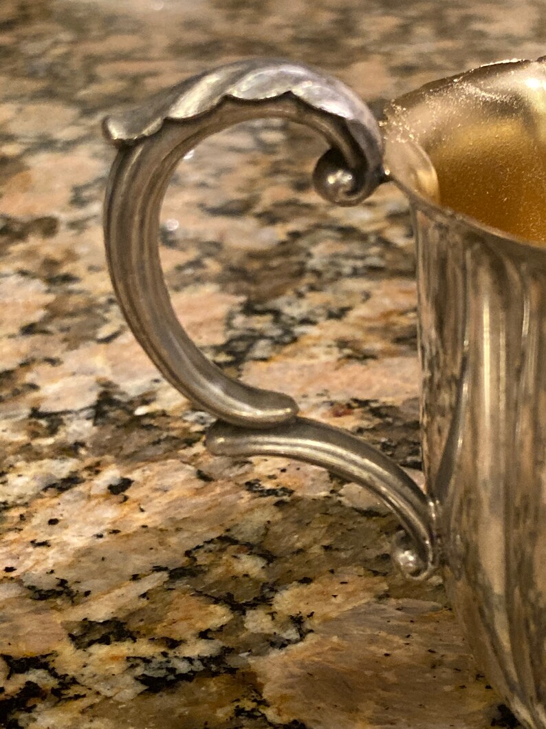 May include: A silver sugar bowl with a decorative handle. The bowl has a fluted design and is made of metal.