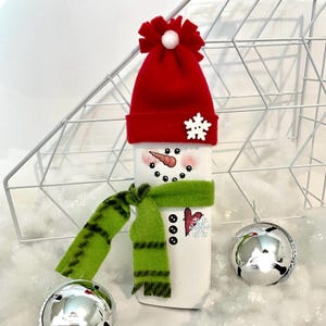 May include: A white snowman-themed bottle cover with a red hat and green scarf. The snowman has a carrot nose, black eyes, and a smiling mouth. A snowflake and heart are on the bottle. Silver ornaments are in the background.