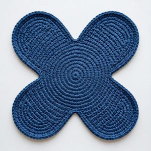 May include: A dark blue crochet coaster or mat featuring a four-lobed, flower-like shape. The item is constructed with concentric circular stitches that radiate from the center, creating a textured, uniform surface throughout the entire piece.