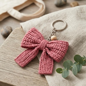May include: A dusty rose crochet bow keyring featuring a textured yarn construction, a central knot detail, and two hanging ribbon ends. The bow is attached to a silver-toned metal split ring by a short chain and a single small, round, light-coloured wooden bead.