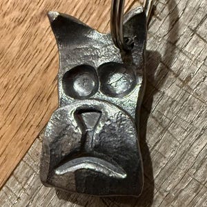 May include: A dark gray metal keychain shaped like a stylized dog head with two circular eyes, a triangular nose, and a curved mouth line. The metal surface has a textured, hammered appearance and includes a circular silver metal split ring at the top.