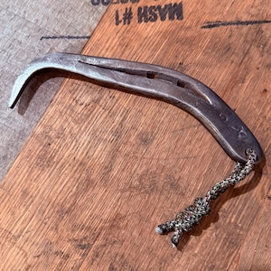 May include: A silver-colored horseshoe-shaped bottle opener with a curved end and a braided cord attached. The opener is on a wooden surface with stamped text.