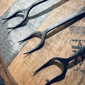 May include: Three hand-forged iron forks with long, slender handles and two sharp, curved tines. The metal has a dark, textured, matte finish with visible hammer marks, showcasing a raw, industrial appearance across the three identical utensils.