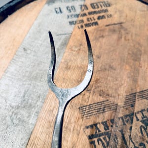 May include: A handcrafted, metal cheese fork with a curved, two-pronged design. The fork is set against a wooden background with visible text and markings, suggesting a rustic or vintage aesthetic. The metal has a slightly aged appearance.