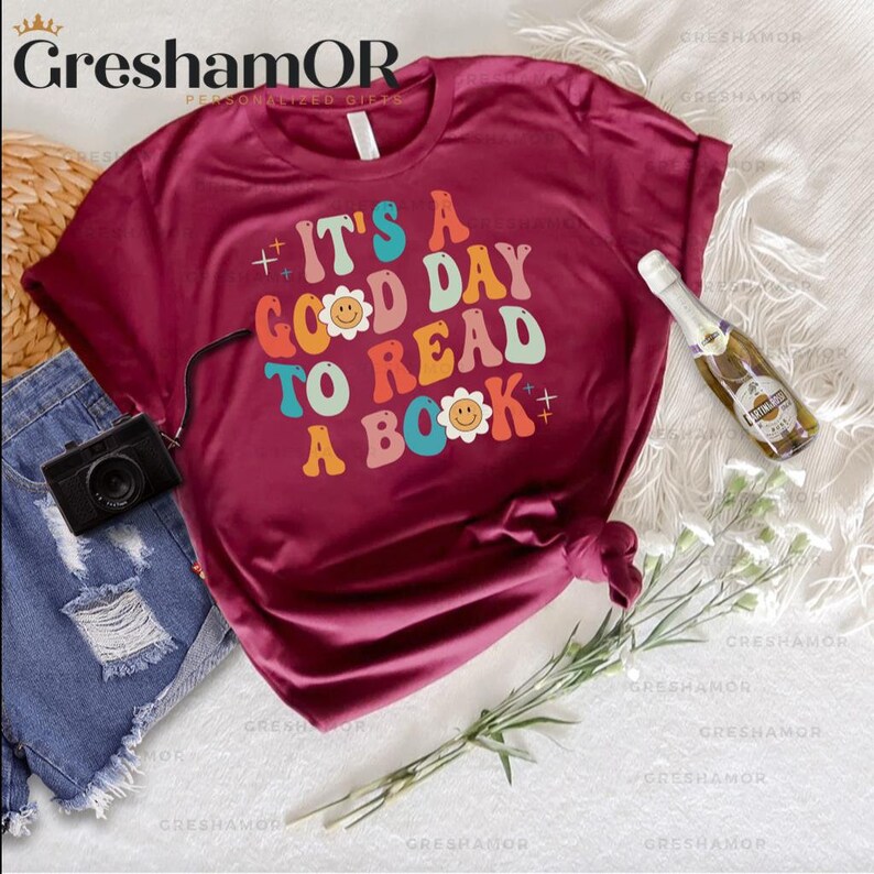 Pode incluir: Uma camiseta vinho com o texto "It's a good day to read a book" em fonte retr&ocirc; colorida com flores com carinhas sorridentes.