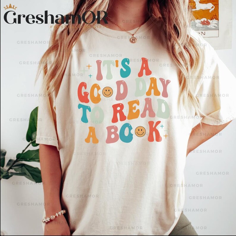Pode incluir: Uma camiseta branca com um gr&aacute;fico colorido que diz "&Eacute; um bom dia para ler um livro". O gr&aacute;fico &eacute; de estilo retr&ocirc; com carinhas sorrindo.