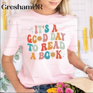 Pode incluir: Uma camiseta rosa com o texto "It's a good day to read a book" em letras coloridas de estilo retr&ocirc;. O texto &eacute; decorado com carinhas sorridentes e flores.