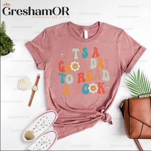 Pode incluir: Uma camiseta rosa com um gr&aacute;fico de estilo retr&ocirc; que diz "&Eacute; um bom dia para ler um livro" com carinhas sorridentes.