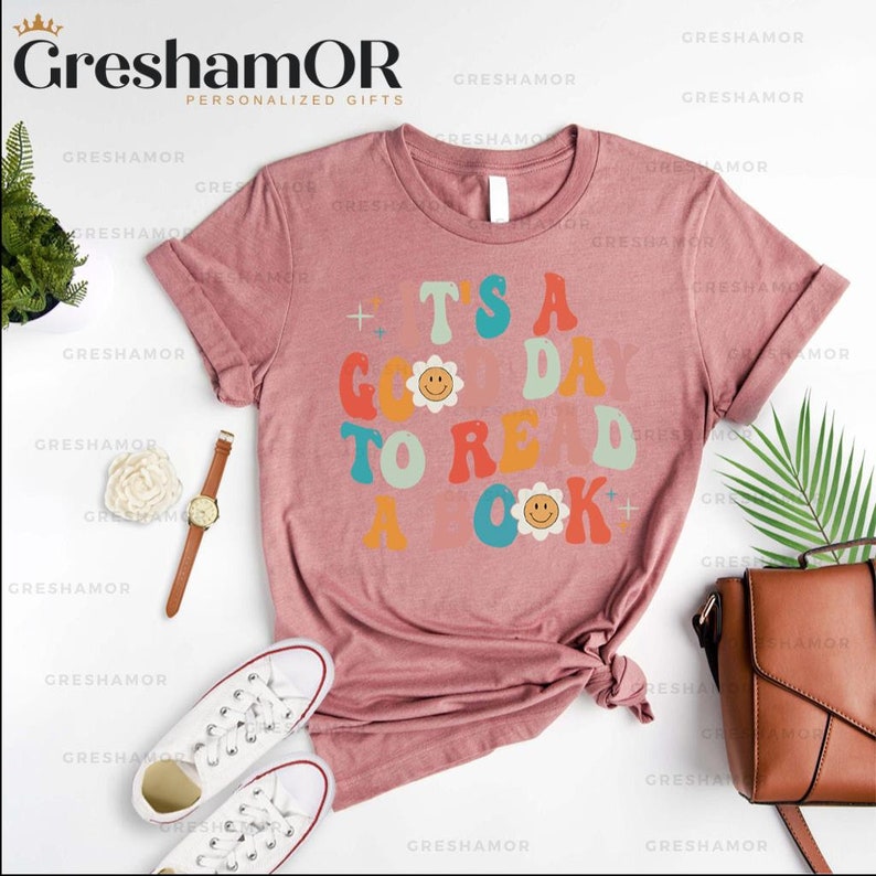Pode incluir: Uma camiseta rosa com um gr&aacute;fico de estilo retr&ocirc; que diz "&Eacute; um bom dia para ler um livro" com carinhas sorridentes.