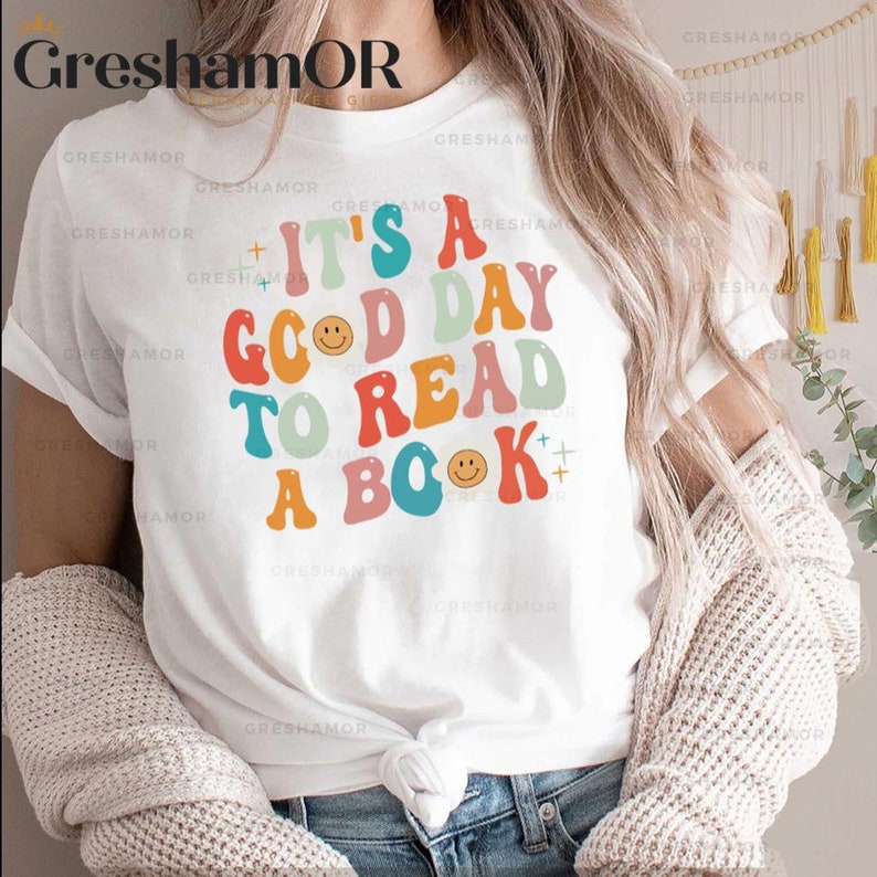 Pode incluir: Camiseta branca com um gr&aacute;fico colorido que diz "It's a good day to read a book". O gr&aacute;fico tem um estilo retr&ocirc; com carinhas sorrindo.