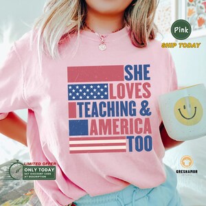 May include: Pink t-shirt with a red, white, and blue American flag design. The text "SHE LOVES TEACHING & AMERICA TOO" is printed on the shirt.