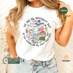 May include: White t-shirt with a colorful graphic of sea creatures in a circle. The text reads "We are all different but in this school we all swim together".