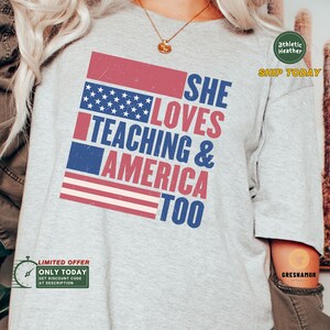 May include: A gray t-shirt with a red, white, and blue American flag design. The text "SHE LOVES TEACHING & AMERICA TOO" is printed on the shirt.