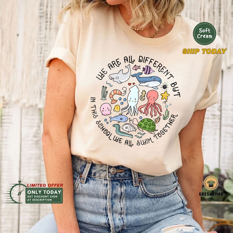 May include: A light beige t-shirt with a circle graphic of colorful cartoon sea creatures. The text around the circle reads "We are all different but in this school we all swim together."