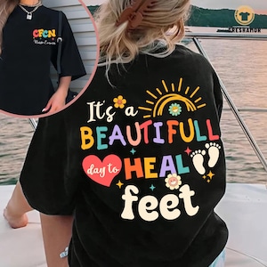 May include: Black t-shirt with a colorful graphic that says "It's a beautiful day to heal feet". The graphic includes a heart, a rainbow, a sun, and footprints.