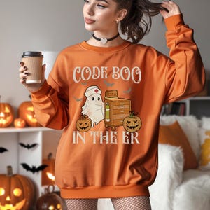 May include: Orange sweatshirt with the text "CODE BOO IN THE ER" and a graphic of a ghost wearing a nurse's hat, a medical cabinet, and jack-o'-lanterns. The sweatshirt is a Halloween-themed design.