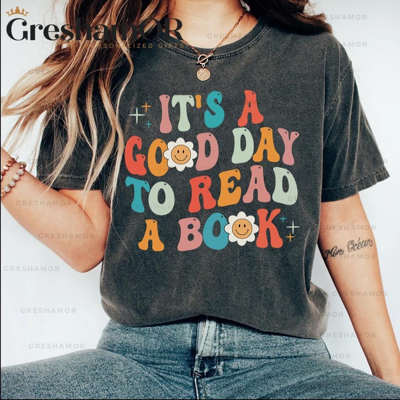 Pode incluir: Uma camiseta cinza escura com um gr&aacute;fico colorido que diz "It's a good day to read a book". O gr&aacute;fico apresenta uma flor com um rosto sorridente no centro de cada palavra.