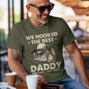 May include: Green t-shirt with a cartoon fish wearing sunglasses and a hat. The text on the shirt reads "We hooked the best * Daddy * Brian Nancy Terry Madison".