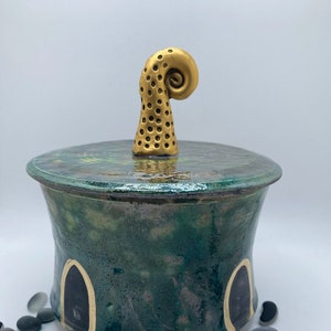 Puede incluir: Un tarro de cerámica con un esmalte verde y dorado. La tapa tiene un pomo dorado en forma de espiral. El tarro está decorado con detalles en negro y dorado. Pequeñas piedras lisas están esparcidas alrededor del tarro.