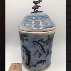 Peut inclure: Un pot en céramique bleu avec un couvercle orné d'une pile de trois sculptures marines argentées. Le pot est décoré d'un motif aquarelle blanc et bleu et de silhouettes noires de différentes créatures marines, notamment des baleines, des dauphins et des requins. Le pot mesure environ 1 pouce de haut.