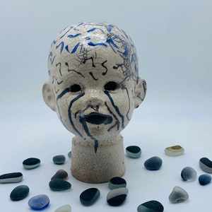 May include: A ceramic doll head with blue and black paint details. The doll head is cracked and has a distressed look. The doll head is sitting on a white surface with small, smooth, gray stones around it.