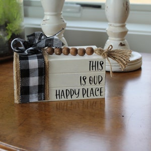 May include: A white wooden block sign with the text "This is our happy place" stacked on top of each other. The sign is decorated with a black and white checkered ribbon, a brown wooden bead garland, and a burlap bow.