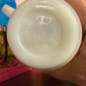 Pu&ograve; includere: Una tazza vintage bianca e opaca con manico. Il fondo della tazza presenta un'area leggermente incassata con testo in rilievo. La tazza &egrave; tenuta da una persona.