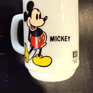Pu&ograve; includere: Tazza in ceramica bianca con un'illustrazione di Topolino. La tazza ha un manico e la parola "MICKEY" stampata in nero. La tazza presenta anche la scritta "PEPSI COLLECTOR SERIES".