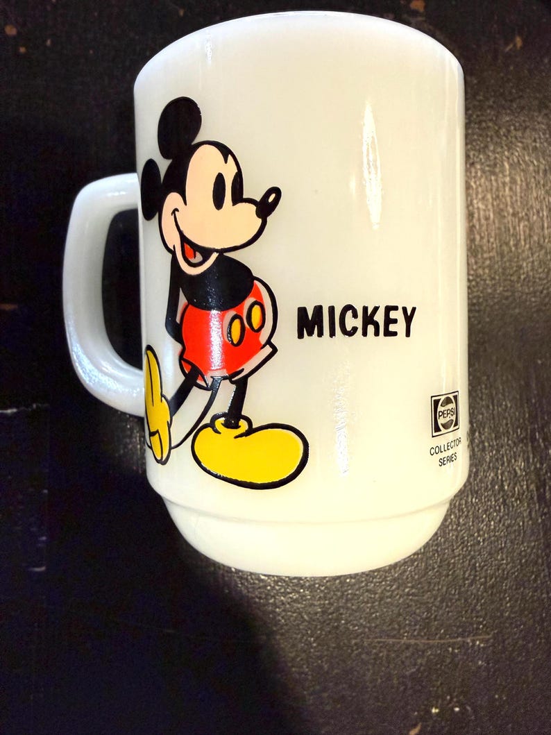 Pu&ograve; includere: Tazza in ceramica bianca con un'illustrazione di Topolino. La tazza ha un manico e la parola "MICKEY" stampata in nero. La tazza presenta anche la scritta "PEPSI COLLECTOR SERIES".