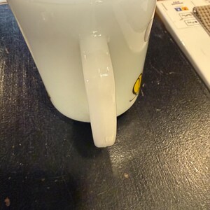 Pu&ograve; includere: Tazza in ceramica bianca di forma quadrata con manico curvo. La tazza ha un disegno giallo sul lato. La tazza &egrave; appoggiata su una superficie scura.