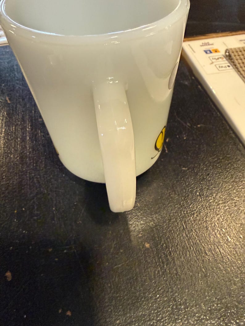 Pu&ograve; includere: Tazza in ceramica bianca di forma quadrata con manico curvo. La tazza ha un disegno giallo sul lato. La tazza &egrave; appoggiata su una superficie scura.