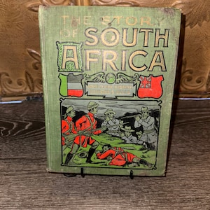 May include: A green book titled "The Story of South Africa" with a gold embossed title and illustrations of soldiers in red uniforms. The book cover features flags of the Netherlands, the Transvaal Republic, and the United Kingdom.