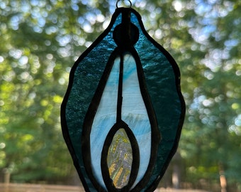 Puede incluir: Una obra de arte en vidriera con forma de vulva estilizada. La pieza presenta tonos de vidrio azul y blanco, con un borde negro. Se suspende mediante una cadena y un anillo, con un fondo verde borroso.