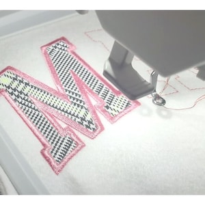 May include: An embroidery machine stitching a pink-outlined letter "M" onto white fabric. The letter has a black, white, and light green plaid pattern. The text "embroidery design" is on the right side of the image.