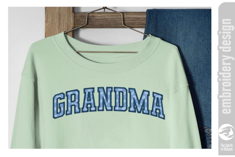May include: A light green sweatshirt with the word "GRANDMA" in blue block letters. The letters have a blue camouflage pattern. The sweatshirt is hanging on a wooden hanger with a pair of blue jeans in the background.