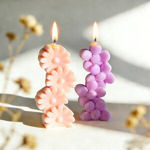 May include: Two lit flower-shaped candles. One is a light peach color, the other is lavender. Each candle is composed of stacked flower blossoms. The flames are yellow. The candles are on a white surface.