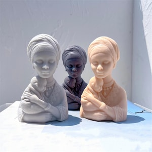May include: Three sculptures of a mother and child in light grey, dark grey, and peach hues. Each mother figure wears a head wrap and cradles a child. The sculptures are detailed and have a calm expression, evoking a sense of peace.