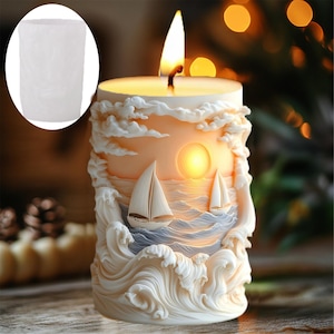 May include: A lit, cylindrical candle with a nautical design. The candle features a sculpted scene of sailboats on the ocean, with waves, clouds, and a setting sun. The candle is ivory-colored and has a small, inset, white, cylindrical container.