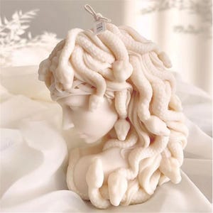 Pode incluir: Uma vela branca esculpida à imagem de Medusa, com cobras como cabelo. A vela é detalhada com traços realistas e é uma peça decorativa única.
