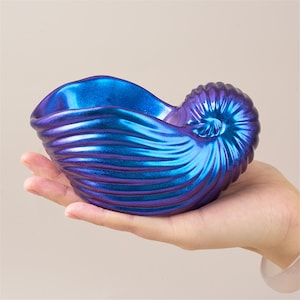 May include: A blue and purple iridescent nautilus shell-shaped bowl. The bowl is held in a hand, showcasing its detailed, ridged exterior. The color shifts from blue to purple, creating a shimmering effect. This decorative piece is ideal for holding small items.