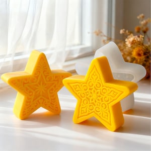 Pode incluir: Dois objetos amarelos em forma de estrela com intrincados desenhos em relevo. Um é uma estrela sólida, o outro é um molde de silicone. As estrelas estão sobre uma superfície branca perto de uma janela com uma cortina transparente e flores secas.