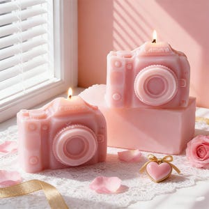 May include: Two pink camera-shaped candles with lit wicks. The candles have a detailed design, including a lens and buttons. The word "Camera" is printed on top of each candle. The candles are on a white lace surface with rose petals and a heart-shaped trinket.