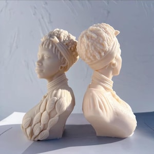 May include: Two off-white busts of women, side by side. Each bust features detailed hairstyles, head wraps, and delicate facial features. One bust has a leaf-like design on the shoulder. The sculptures are set against a light background.
