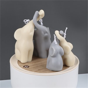 May include: A selection of elephant-shaped candles in shades of grey and cream, displayed on a wooden-effect platform. Each candle has a wick and sits on a white base. These candles are a decorative item.