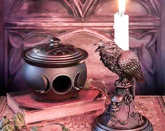 May include: A decorative gothic-style scene featuring a raven candle holder with a lit white candle, a cauldron with crescent moon designs, and a closed book. The raven candle holder has a skull base. The items are in shades of brown and purple.