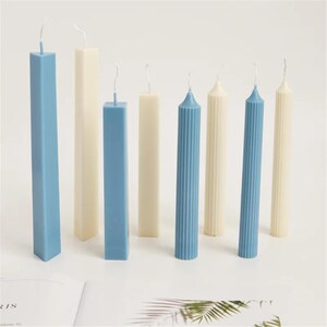 May include: An assortment of candles in various shapes and colors. The candles include tall, rectangular candles in light blue and cream, and fluted, cylindrical candles in blue and cream. The wicks are visible at the top.